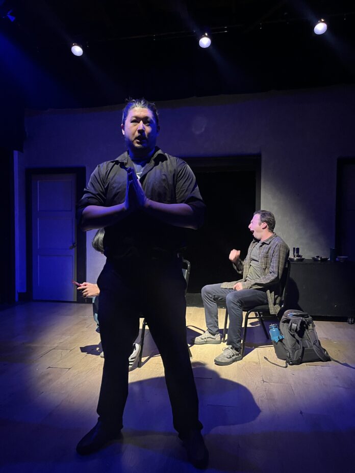 My Tiget Actor stands center stage in prayer pose during My Tiger at the Brickhouse Theatre, with two other performers seated in the background.