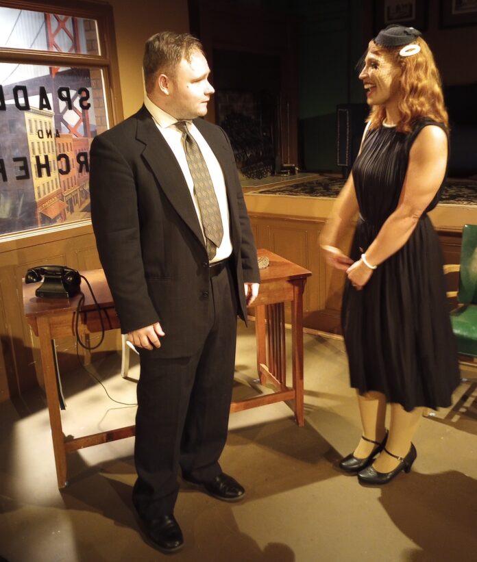 Scene from ELATE Theatre’s The Maltese Falcon — a tense moment as a man unwraps a mysterious package while a woman looks on and another man lies motionless on the floor.