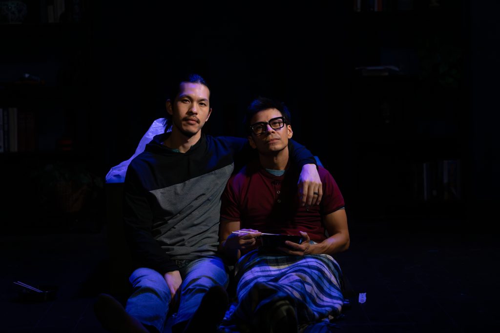 Luca and Uri seated together in dramatic stage lighting