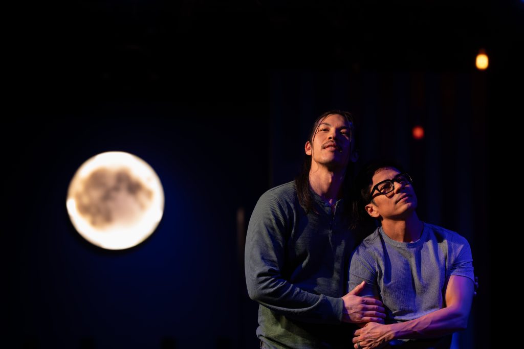 Luca and Uri standing together under stage moonlight