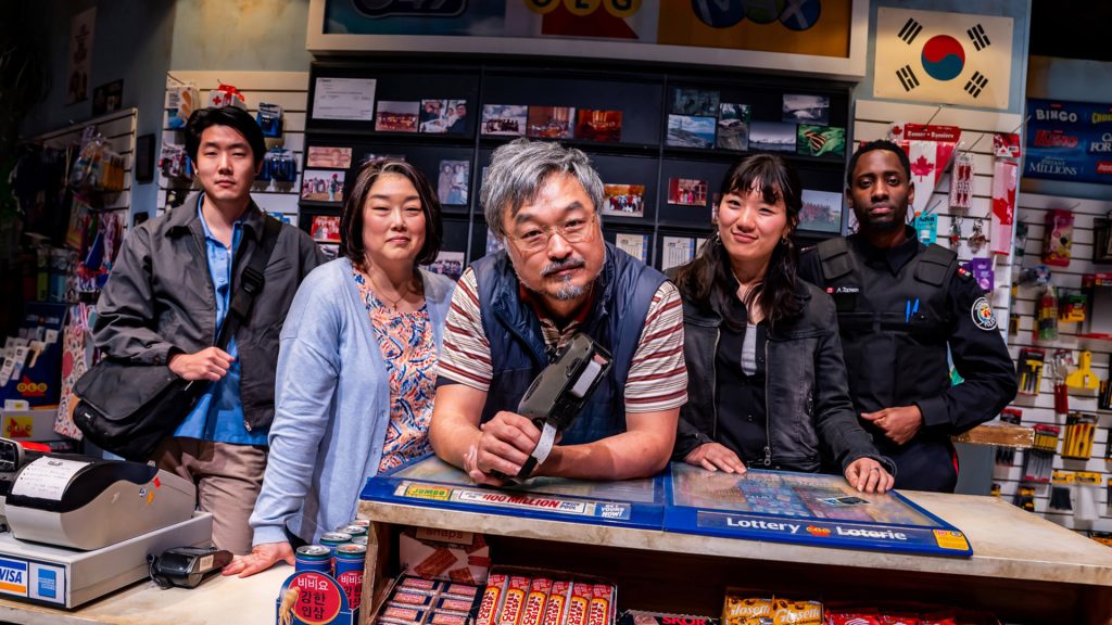 Full cast of Kim’s Convenience behind a convenience store counter, surrounded by snacks and lottery displays