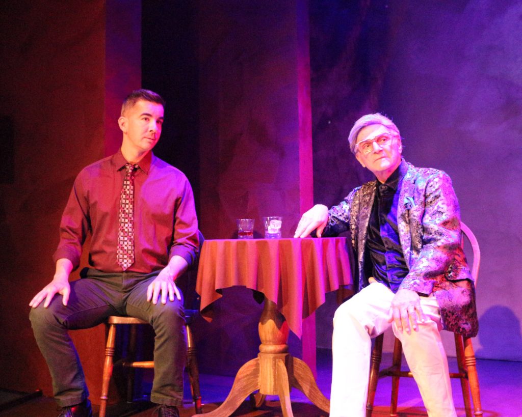 Danny Lee Gomez as Tim sits across from Tony Abatemarco as Spencer at a small table with drinks in Hell Mouth at The Road Theatre in NoHo Arts District