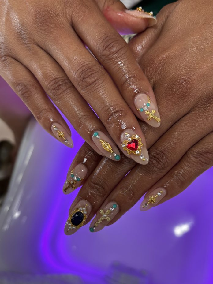Intricate nail art with gold charms, gemstones, crosses, and turquoise accents on almond-shaped nude nails at Beauty Haus NoHo