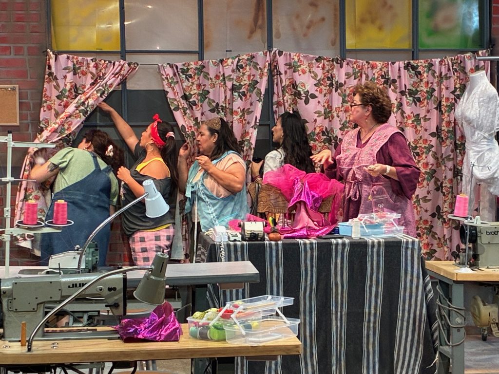 Real Women Have Curves stage production showing group of women in garment factory setting,peeking out a window to see if ICE is there.