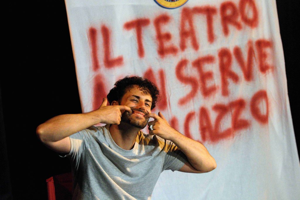 Danilo Napoli performing in The Show Has Been Cancelled with a forced smile in front of a banner reading “Il Teatro al Serve Cazzo” during the Hollywood Fringe Festival solo show.