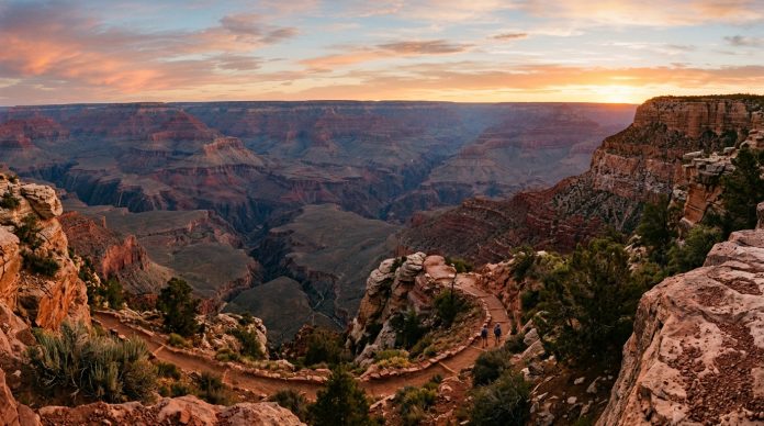 Sunrise view of the Grand Canyon with layered red rock cliffs, winding canyon trails, rugged desert terrain, and expansive panoramic vistas beneath a glowing orange and pink sky.