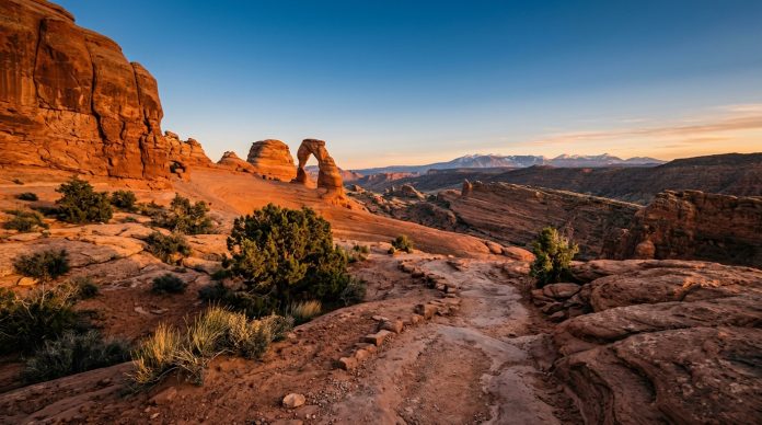 Delicate Arch in Arches National Park at sunset with glowing red sandstone cliffs, desert trails, rugged canyon landscapes, and distant mountain views beneath a clear blue sky.