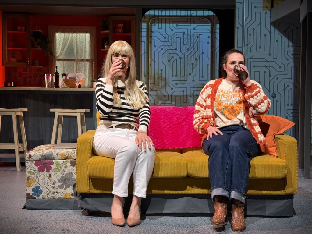 Two performers sitting on couch during comedic scene in Millennials Are Killing Musicals