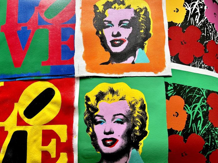 (Warhol flat lay) Artist Eric Doeringer's bootleg paintings of Andy Warhol works including Marilyn Monroe silkscreens, Flowers, and LOVE sculpture recreations laid out on a green surface.