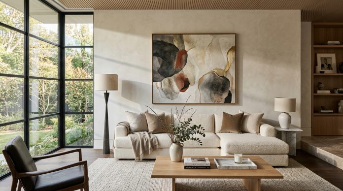 Modern luxury living room with oversized abstract wall art above a neutral sectional sofa, warm natural light, minimalist decor, textured fabrics, and floor-to-ceiling windows overlooking a landscaped garden.
