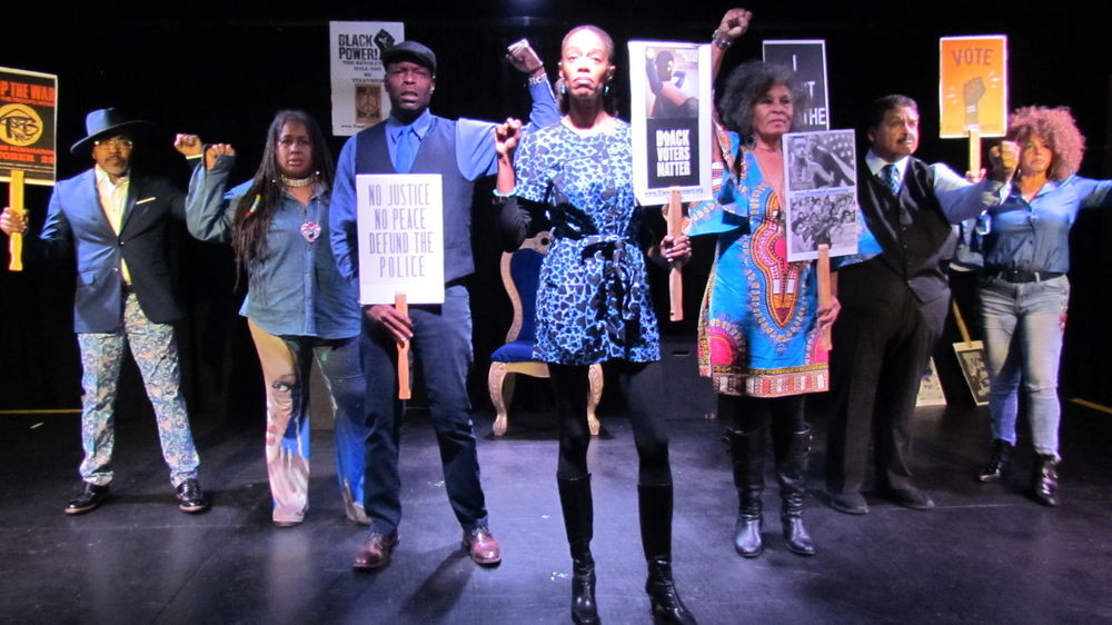 Cast members of Gil Scott-Heron Bluesology hold protest signs and raise their fists during a powerful spoken word performance exploring civil rights, social justice, activism, and the enduring legacy of Gil Scott-Heron.