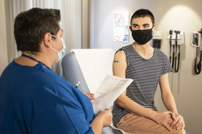 Masked patient speaking with a healthcare worker during a medical appointment in a clinic exam room, representing respiratory health, workplace exposure, and lung cancer awareness.