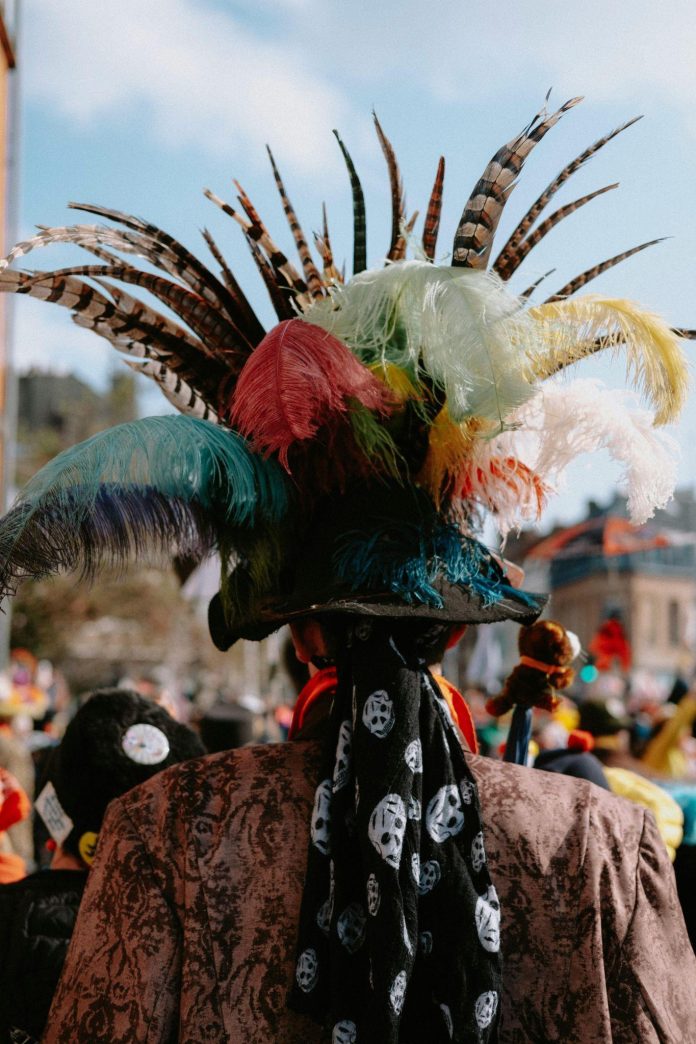 Person in elaborate feathered costume walking through the crowded Edinburgh Festival Fringe with colorful street performers and historic city buildings in the background.