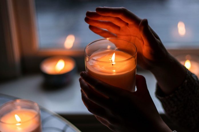 Hands holding a lit candle beside warm glowing candles in a relaxing nighttime home atmosphere