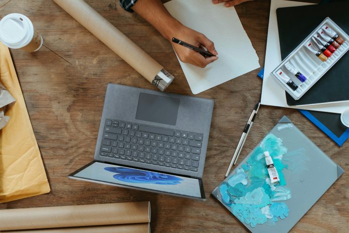 Artist workspace with laptop, sketchbook, paint tubes, and canvas used for creating and selling art on social media