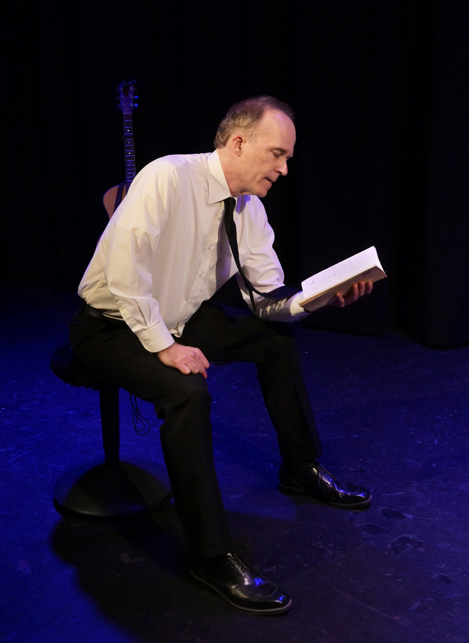 Stefan Marks sits on a stool reading from a notebook during a performance of My One Woman Show (The Search for a Truthful Moment) at the 2026 Hollywood Fringe Festival.