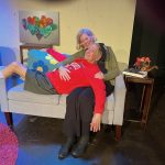 Act Your Age cast members share a tender moment on a living room set at Two Roads Theatre.