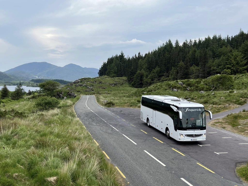Fitzpatrick Coaches tour bus traveling through the scenic countryside of County Kerry surrounded by forests, mountains, and winding roads.