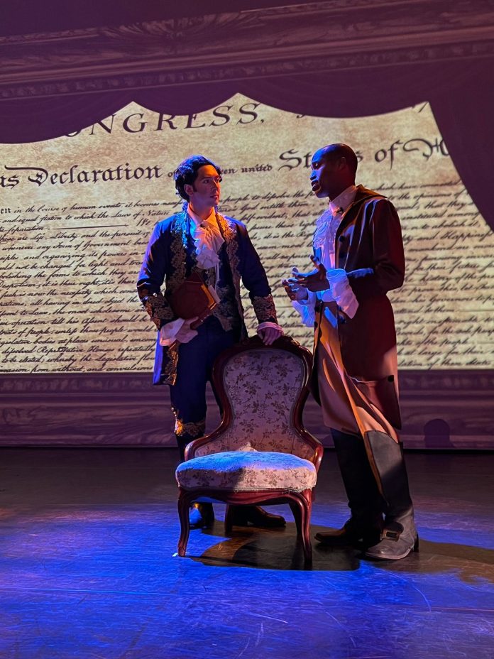 Period-costumed actors perform in The Fighting Quaker at El Portal Theatre, standing before a projection of the Declaration of Independence as Timothy Matlack's role in American history comes to life.