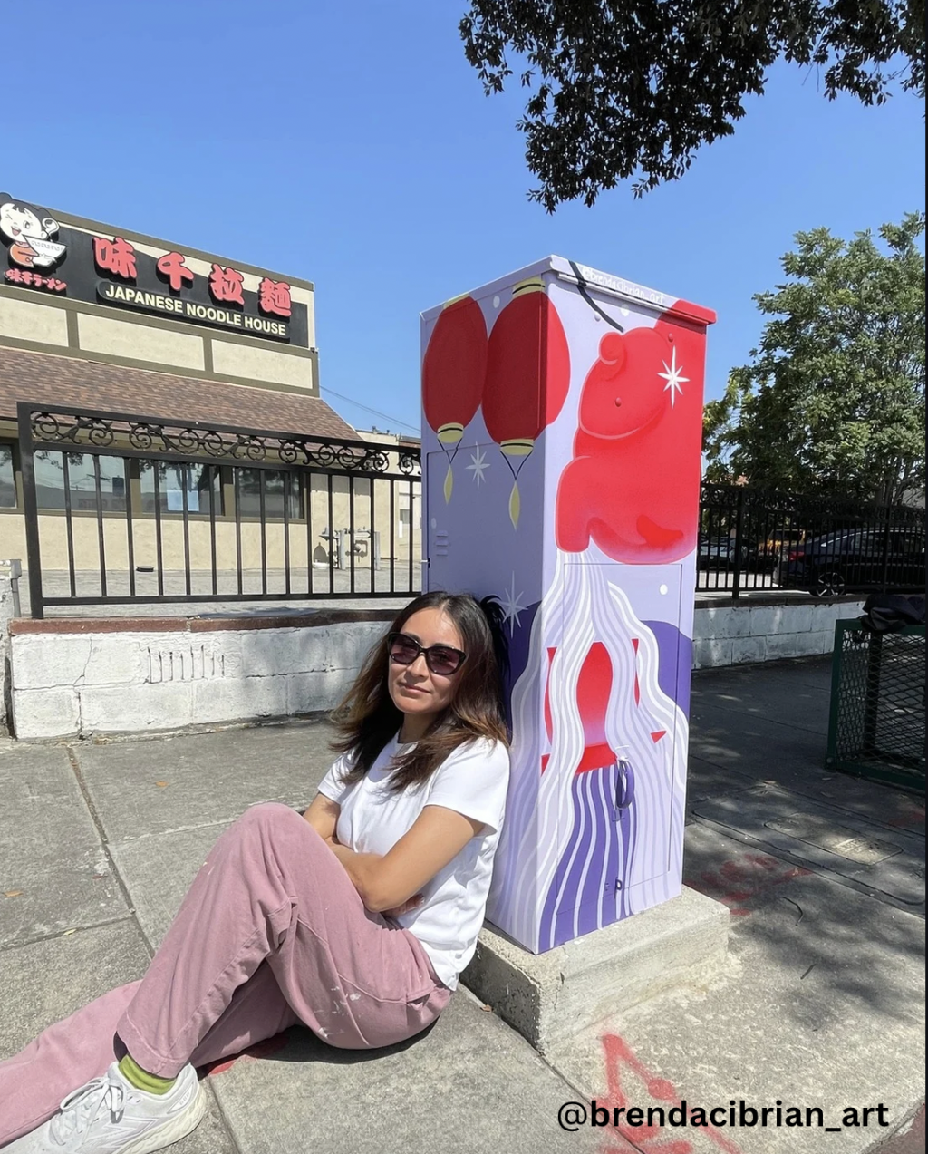 Artist @brendacibrian_art poses beside a completed utility box artwork featuring lanterns and flowing abstract shapes.