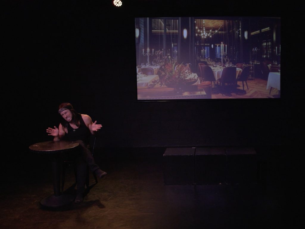 McKinnon Powell performs a scene from 6 out of 10 at the 2026 Hollywood Fringe Festival with a gym image projected behind him on stage.