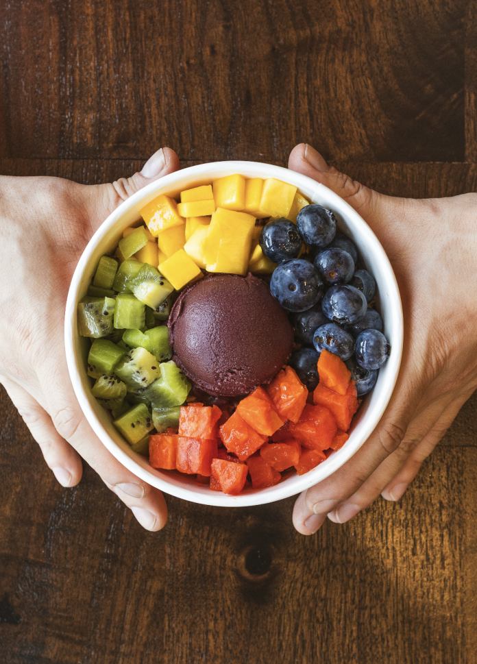 Hands holding a colorful açaí bowl with kiwi, mango, blueberries, papaya, and a scoop of authentic Brazilian açaí.