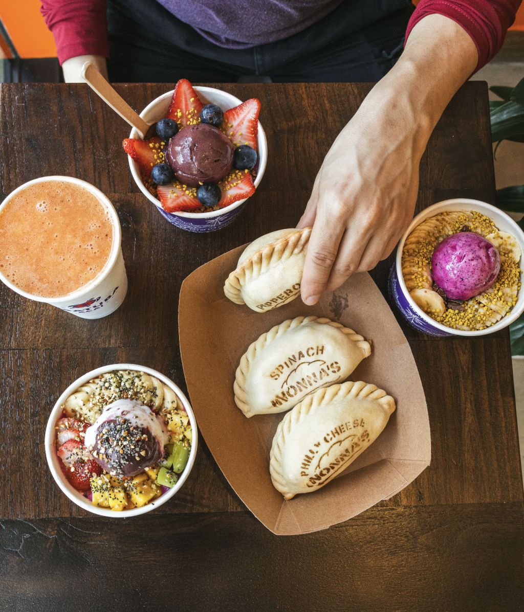 Variety of authentic Brazilian açaí bowls, freshly baked Argentine empanadas, and fresh fruit smoothies from Uba Tuba NoHo displayed together on a wooden table.