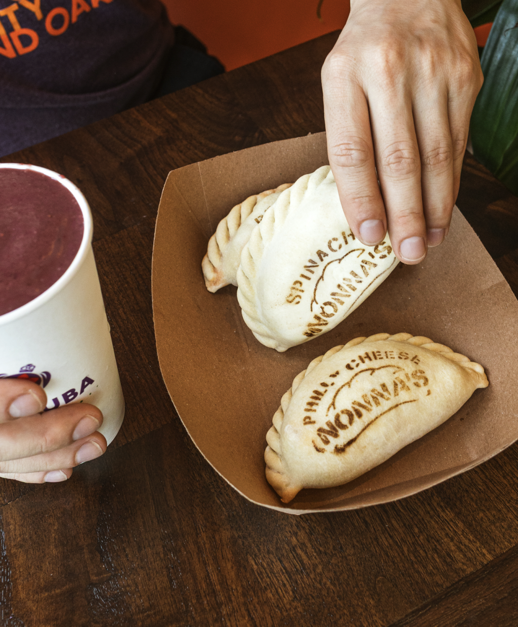 Hand reaching for a freshly baked Argentine empanada with another empanada and a smoothie on the table.