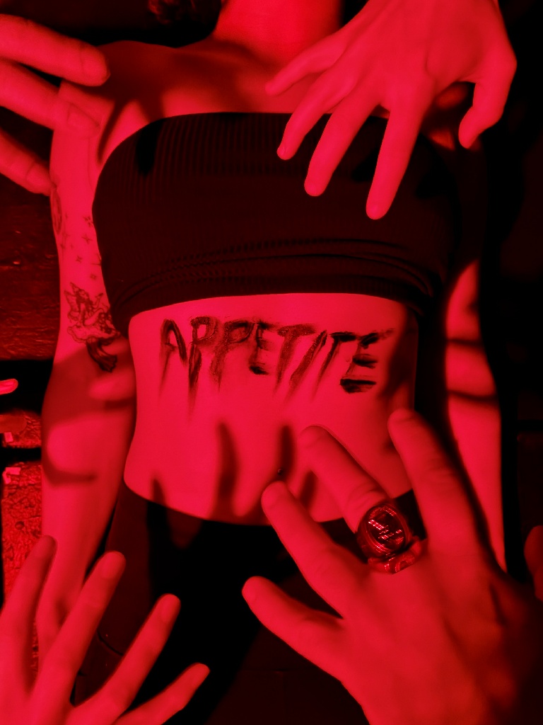 Performers surround a cast member with "APPETITE" written across their torso under eerie red lighting in Zombie Joe's Underground Theatre Group's horror production.