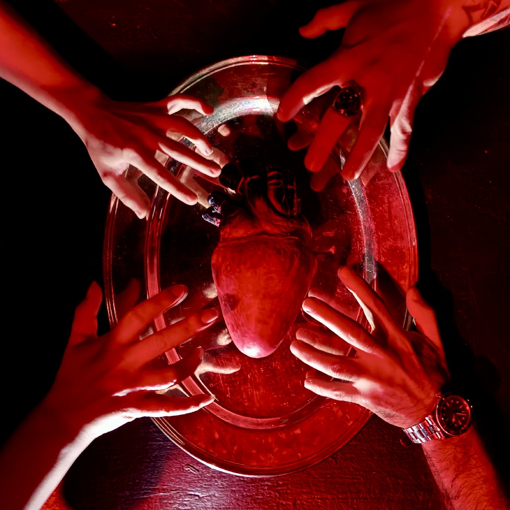 Hands reaching toward a realistic human heart prop under red lighting in a scene from APPETITE at Zombie Joe's Underground Theatre Group.