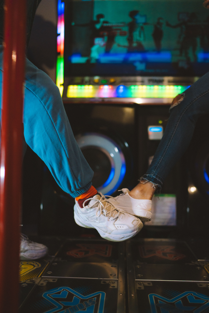Two people playing a dance arcade game on illuminated dance pads, wearing sneakers while following choreography on the game screen.