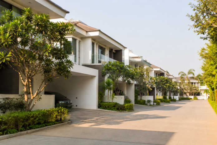 ChatGPT Image Aug 7, 2026, 05_22_42 PM Modern luxury villa community in Hyderabad featuring contemporary white homes with landscaped gardens, tree-lined streets, covered parking, and a quiet residential setting on a sunny day.