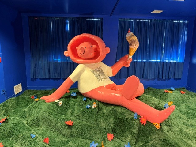Oversized cartoon figure relaxing on artificial grass while holding an ice cream cone in an immersive art installation.
