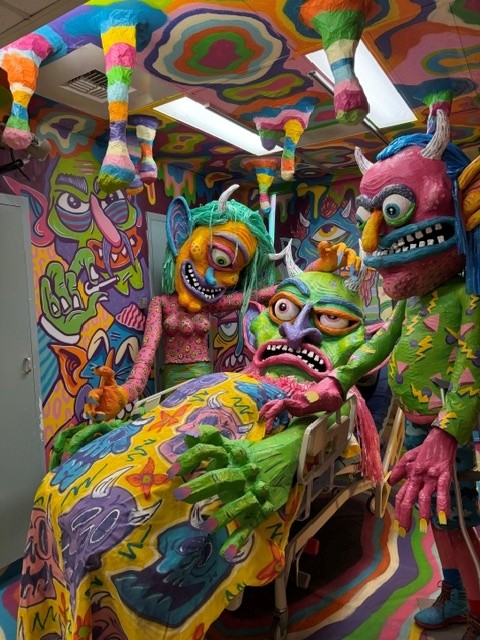 Colorful monster-filled immersive installation with oversized sculptural creatures gathered around a hospital bed.