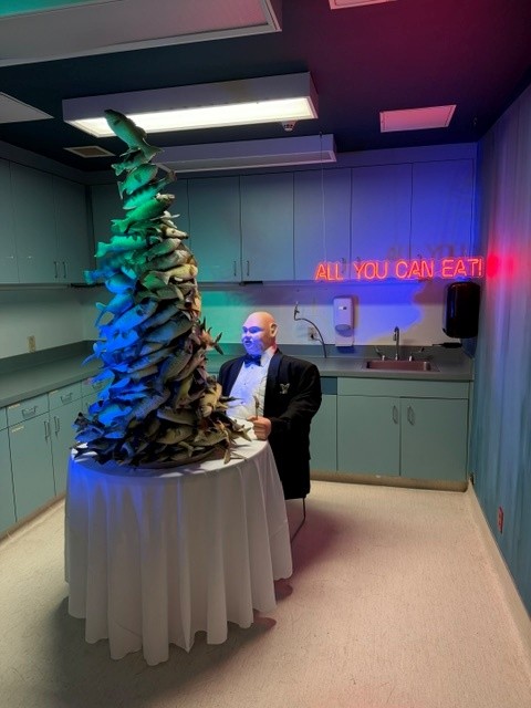 Surreal dining room installation featuring a towering fish sculpture, seated mannequin, and neon "ALL YOU CAN EAT" sign.