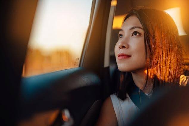 A woman wearing a seatbelt looks out the window from the back seat of a car during golden hour, suggesting safe and comfortable travel.