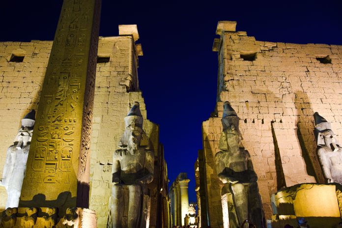 Ancient statues, obelisk and towering stone walls illuminated at Luxor Temple in Egypt at night.