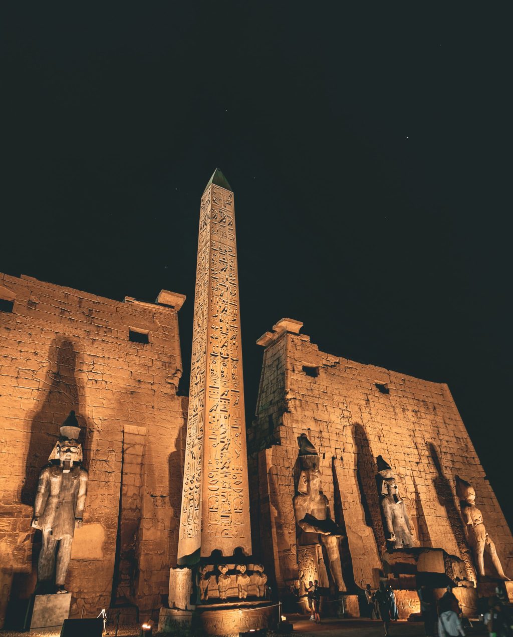 Illuminated obelisk and colossal pharaoh statues at Luxor Temple in Egypt under the night sky.
