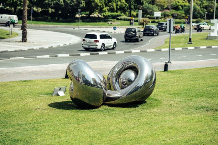 Abstract stainless-steel sculpture on a landscaped lawn in Dubai, reflecting the surrounding cityscape.