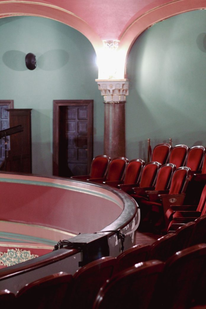 Historic theatre interior with rows of red seats and an ornate curved balcony.