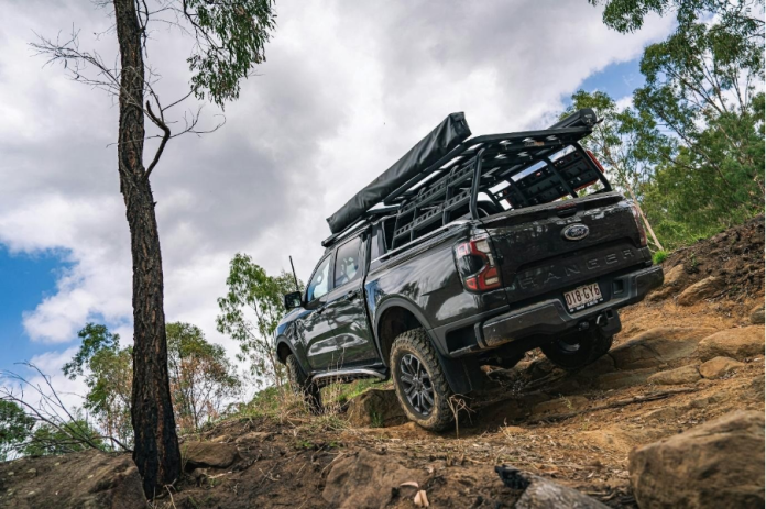 image Black Ford Ranger with a roof rack and overlanding accessories driving off-road through a forest, built for camping and adventure.