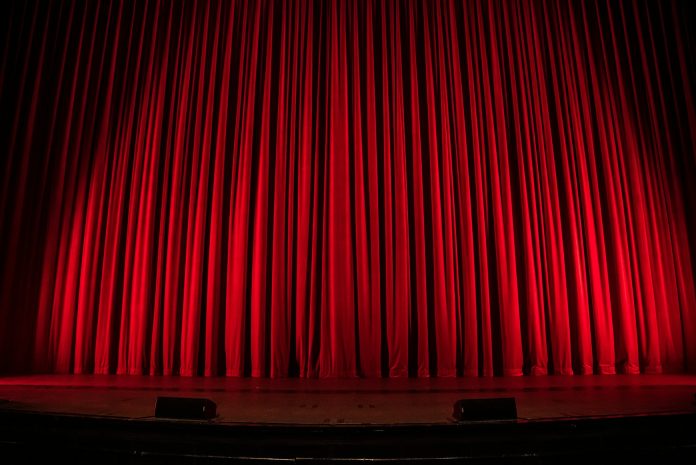 Closed red velvet theater curtains illuminated by stage lights before a live performance, with the empty stage and footlights visible, creating a classic theatrical atmosphere.