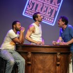 Three performers from Texas Sodomy gathered around a bar beneath a projected Pacific Street Houston, Texas sign.