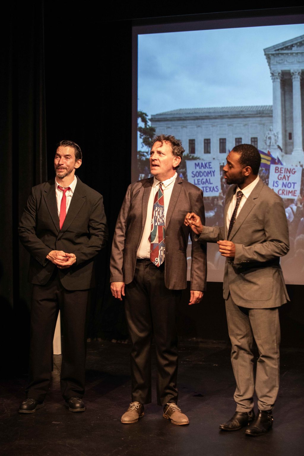 Three performers from Texas Sodomy onstage with a projected image of the U.S. Supreme Court and gay rights protest signs.