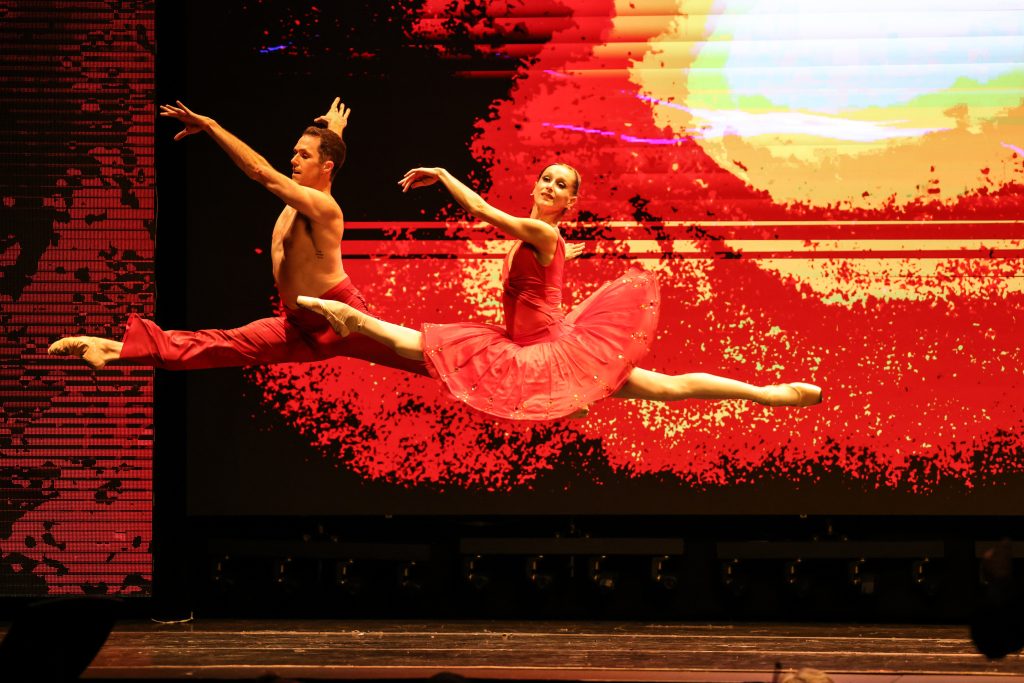 Luminario Ballet dancers performing synchronized leaps against a vivid red stage backdrop.