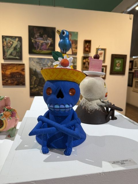 Blue skeleton sculpture topped with fruit and a small bird, displayed among other works at Laluzapalooza 2026.