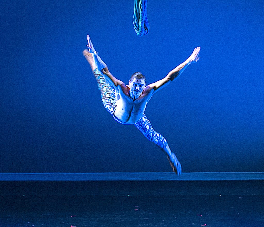 Luminario Ballet dancer performing a dramatic airborne leap under blue stage lighting.
