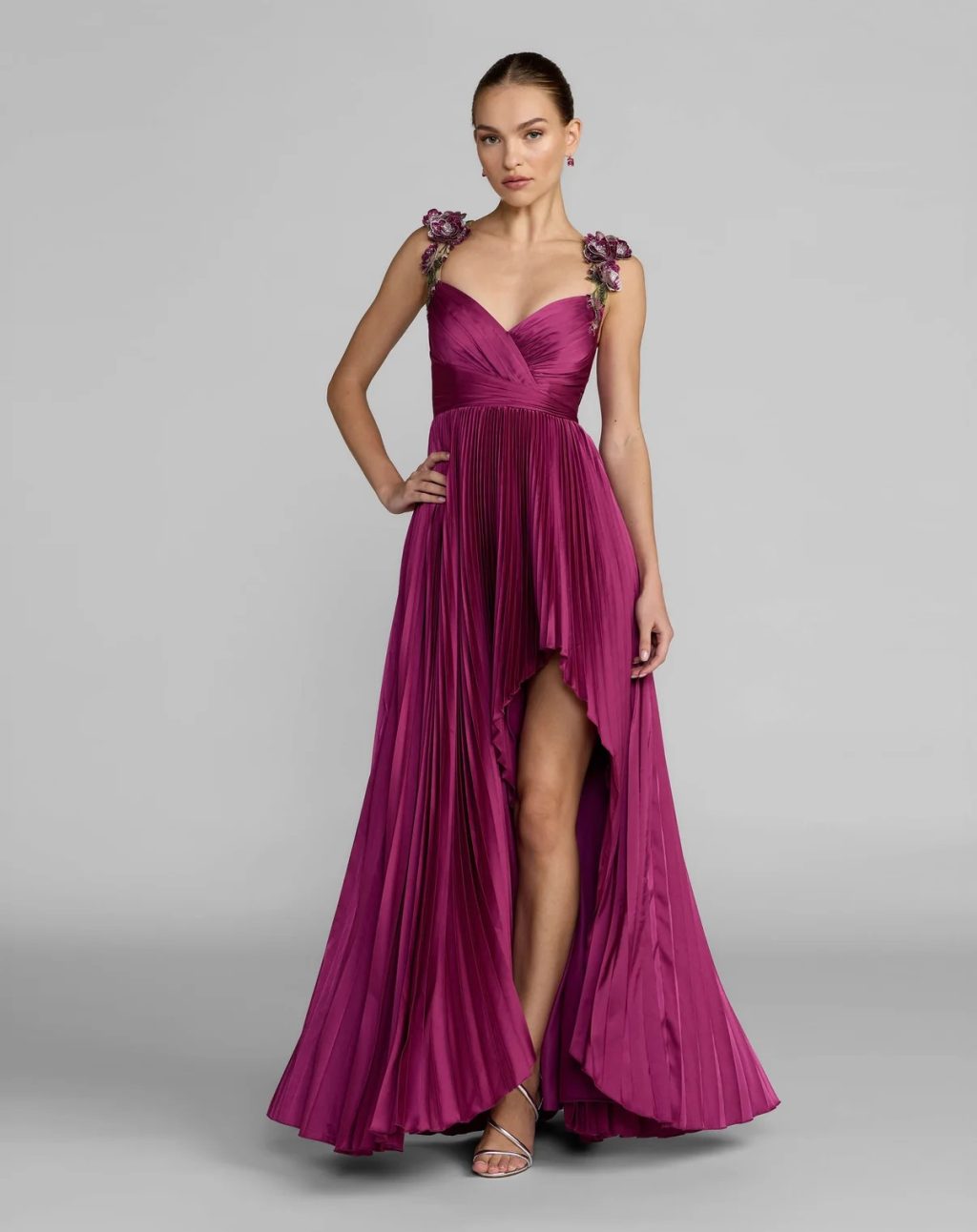 Woman modeling a dramatic magenta evening dress with floral shoulder details and a high-low pleated skirt.