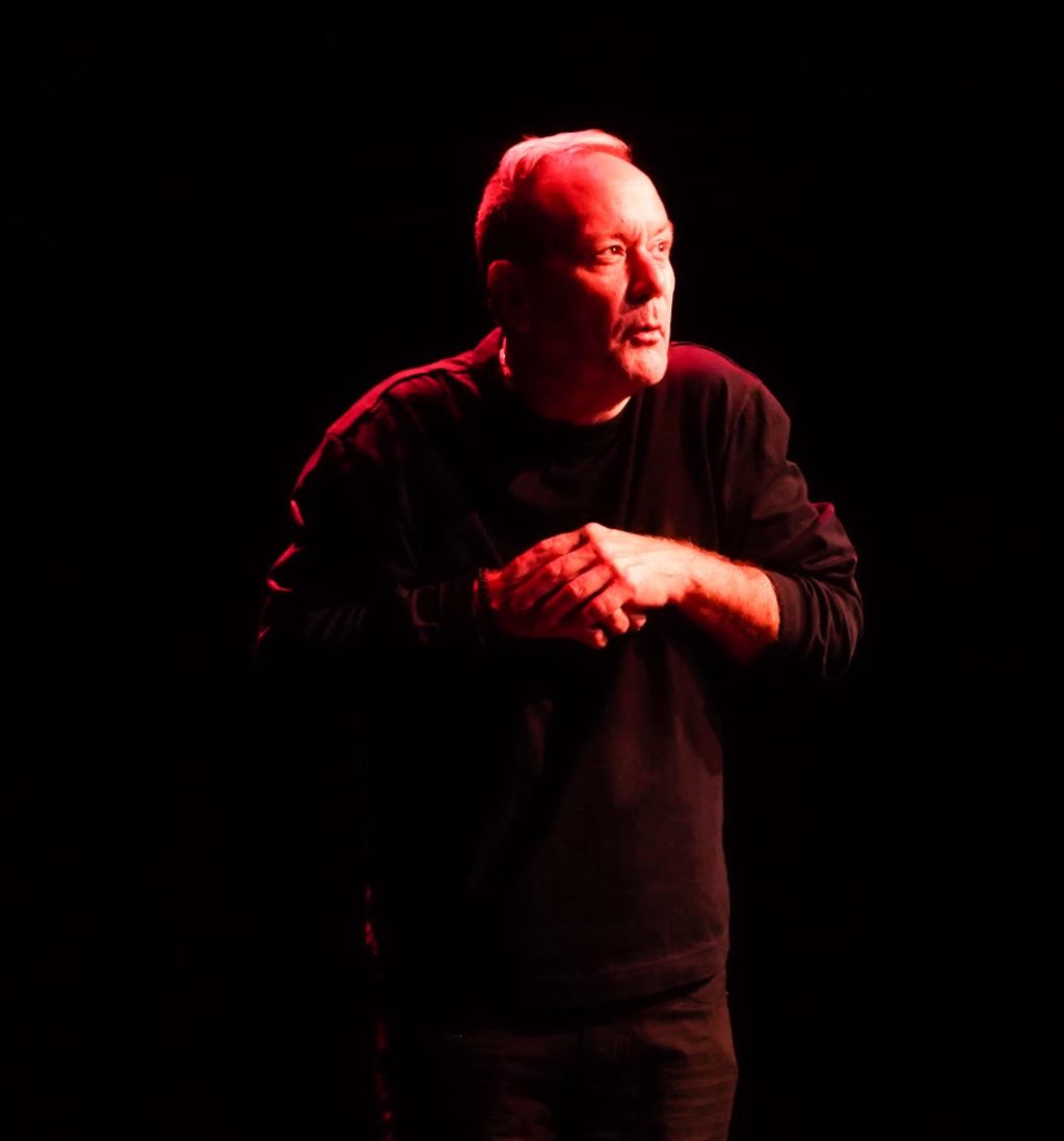Dan Ruth performing onstage under dramatic red lighting during a solo theatre performance. Photo by Tolga Reis.