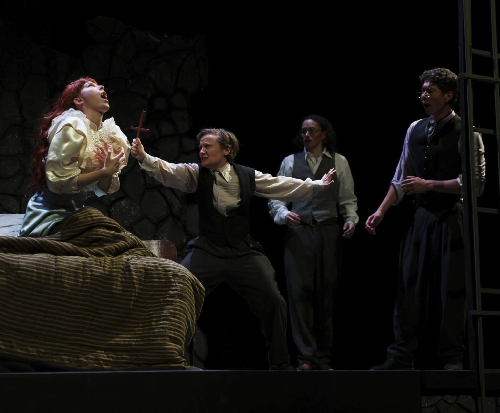 A man holds up a cross toward a distressed woman on a bed as two others look on in Pierce College Theatre's production of Dracula.